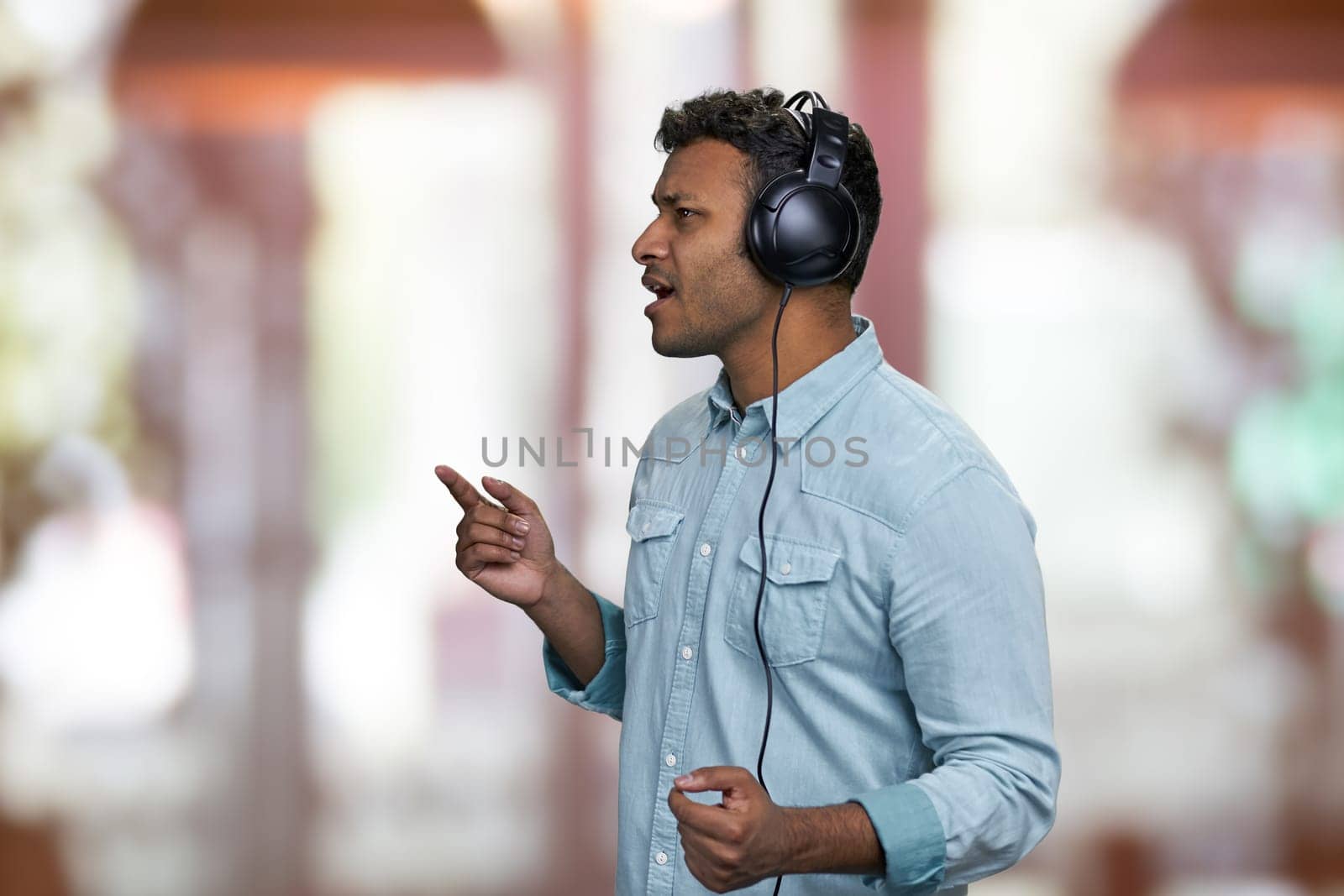 Handsome young man wearing blue shirt listening to music with headphones. Interior blur background.