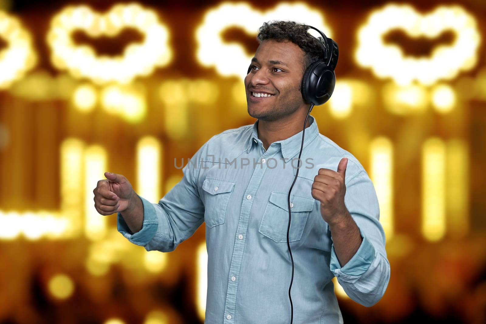 Handsome young man is dancing while listening music with headphones. Golden bokeh lights in the background.