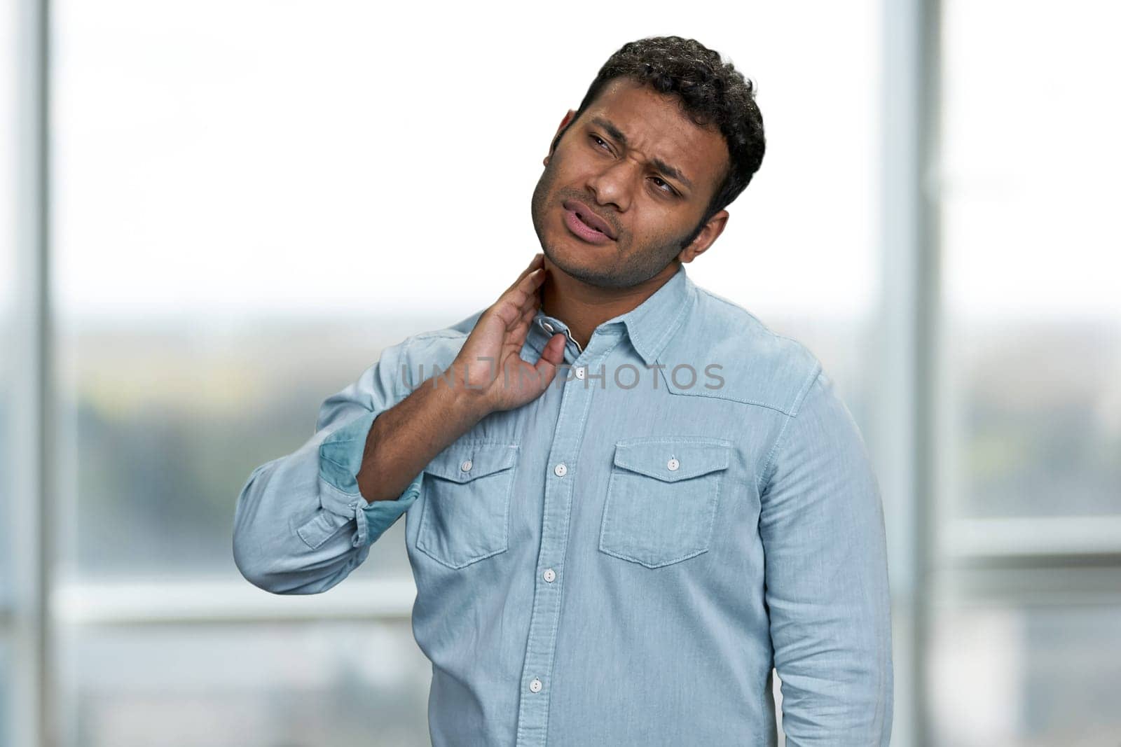 Young indian man suffering from neck pain on blur interior background. People and healthcare concept.