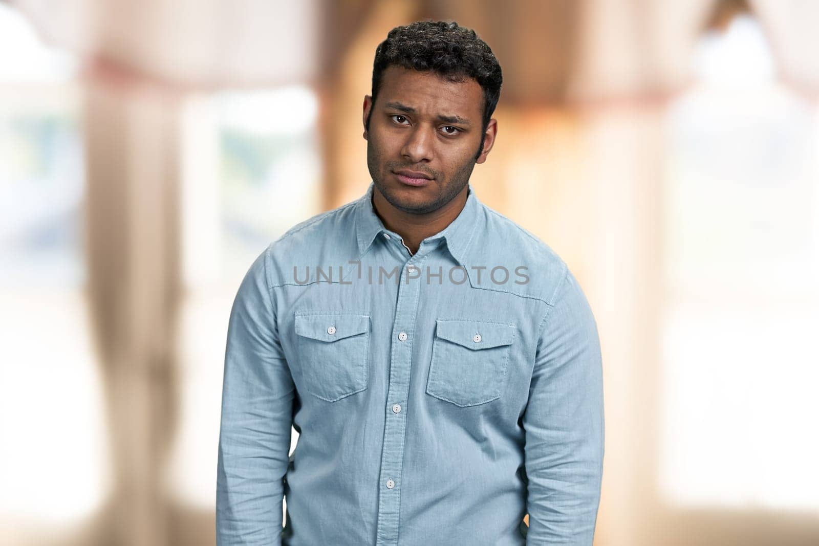 Portrait of upset young indian man looking at camera. Disappointed guy on blur interior background.