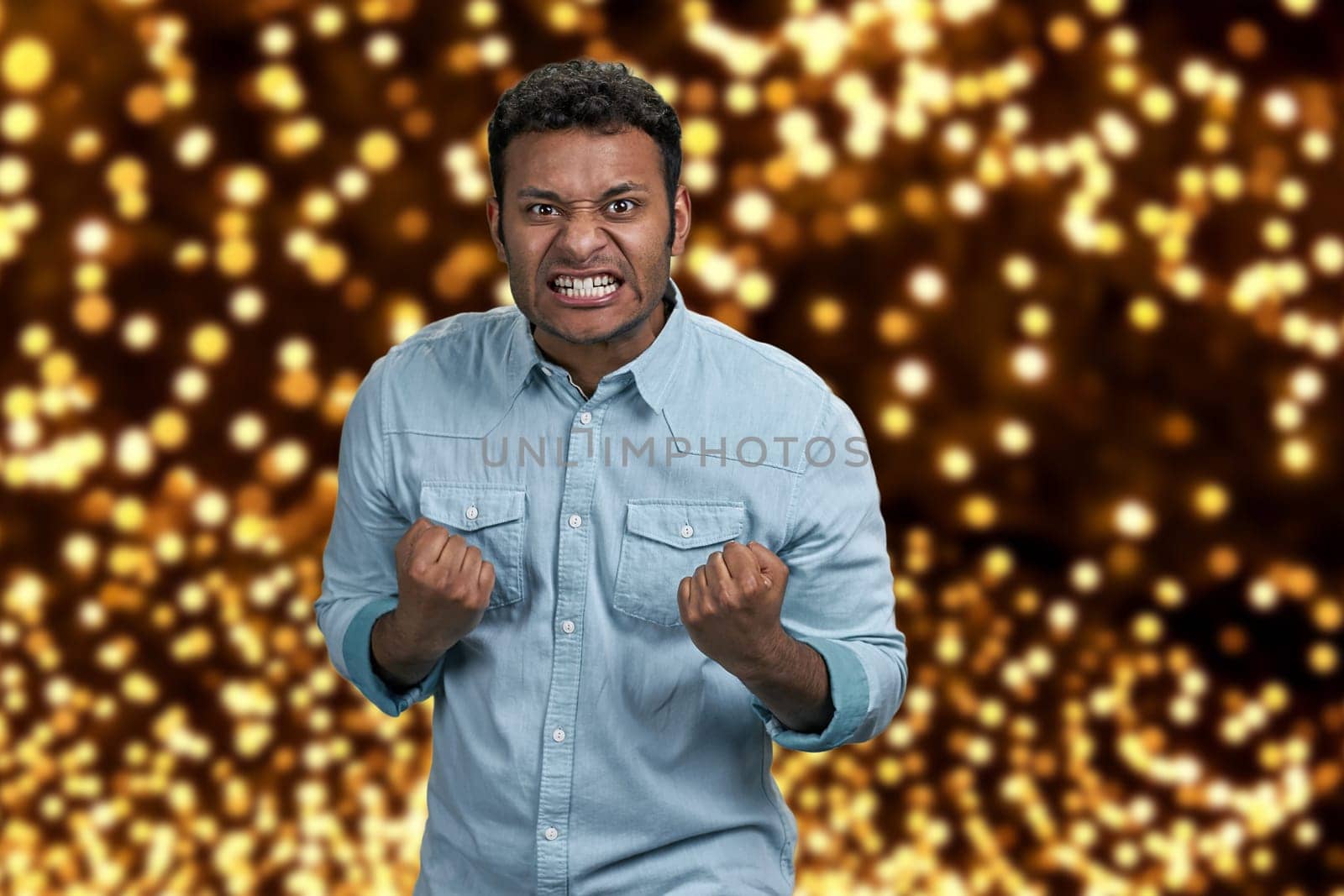 Angry aggressive guy grimacing and shaking fists. Festive bokeh background. Human negative expressions.