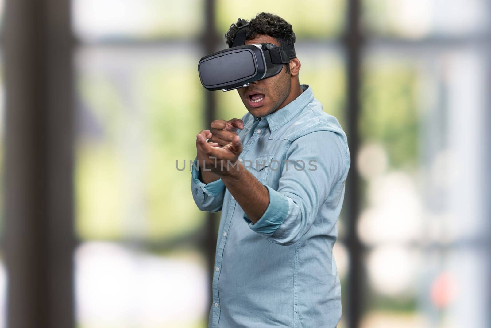 Enthusiastic man in VR glasses simulating shooting with virtual gun. Interior blur background.