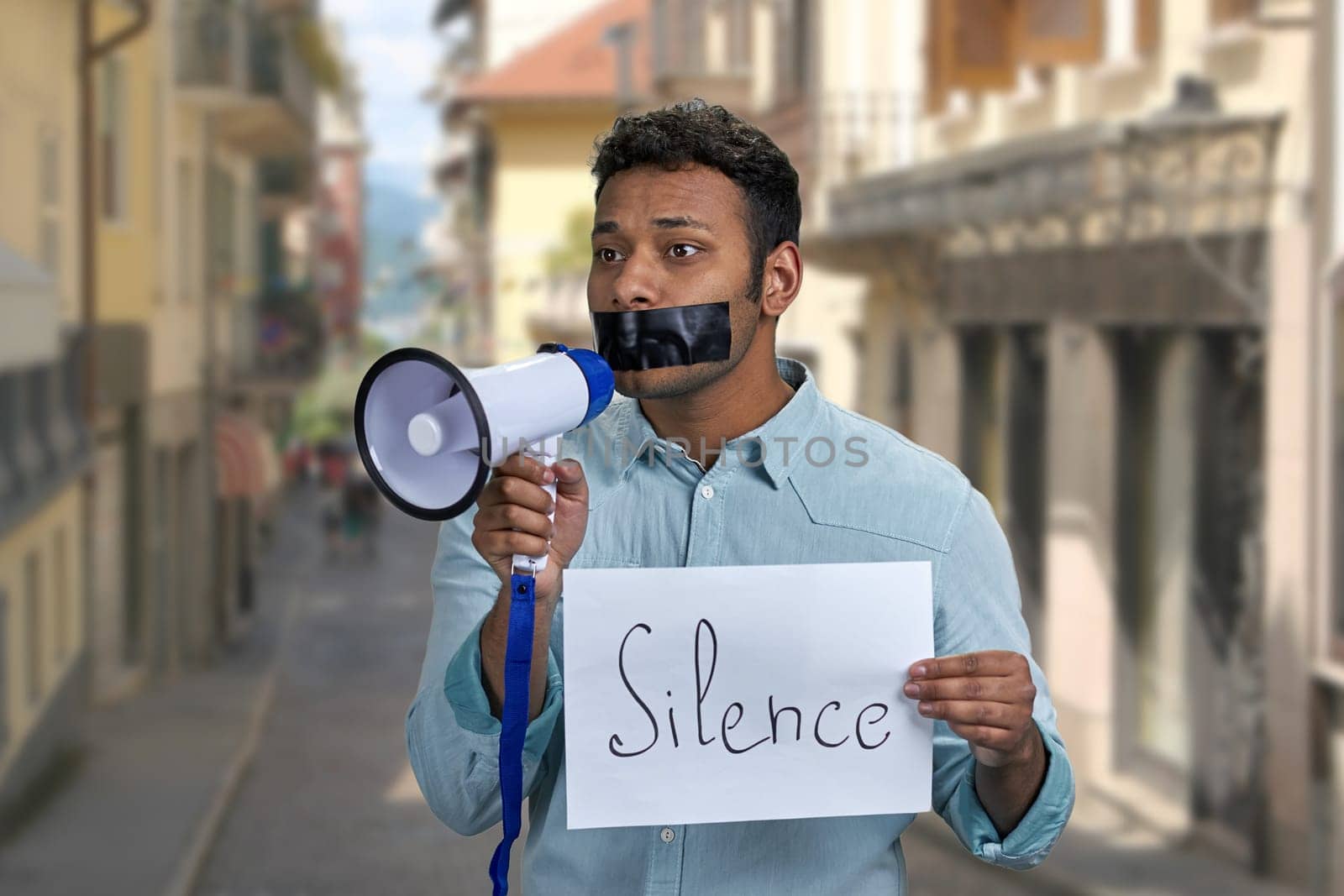 Young indian man with taped mouth unable to speak into megaphone. Censorship concept.