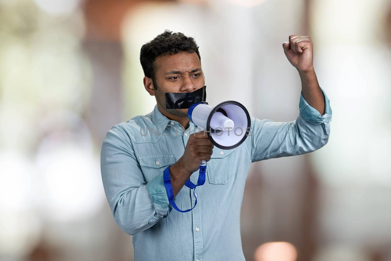 Young man acivist with taped mouth trying to speak into megaphone. Abstract bokeh background.