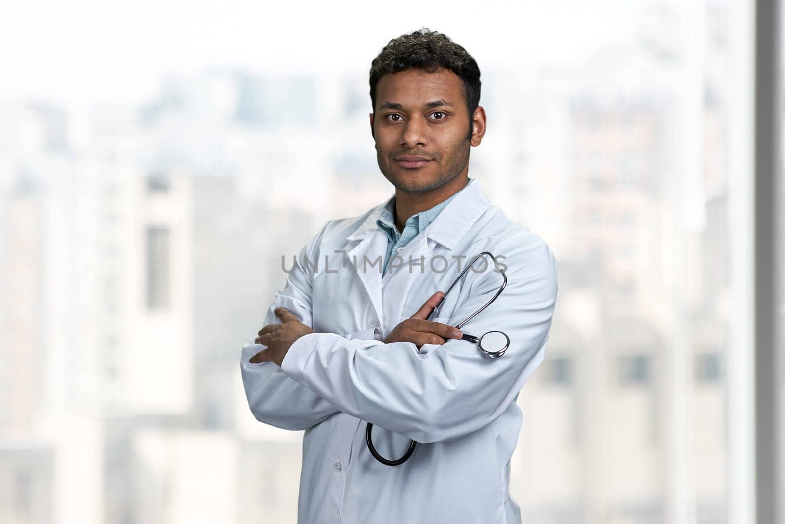 Young confident indian doctor with stethoscope looking at camera standing with arms crossed. People, medicine and occupation concept.