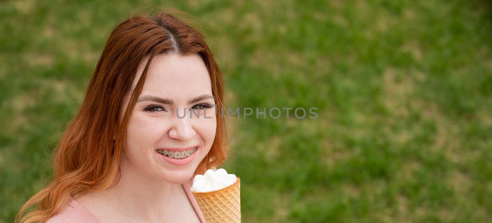 Young beautiful red-haired woman smiling with braces and eating an ice cream cone outdoors. Widescreen. by mrwed54