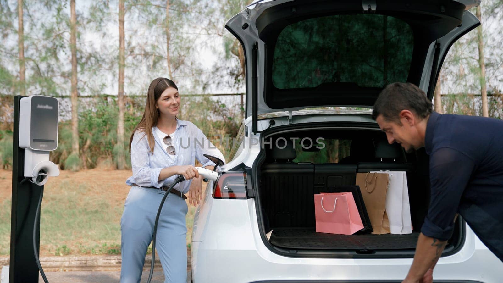 Couple recharge their EV car with electric battery charging station at rest stop during their road trip travel during autumnal season to national park. Relaxing holiday with eco friendly car. Exalt