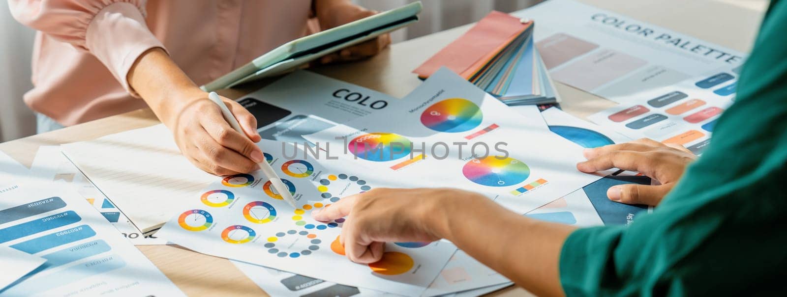 Skilled architect searching data from tablet while selecting an appropriate color from color wheel at table with color palette and document scatter around. Creative design concept. Variegated.