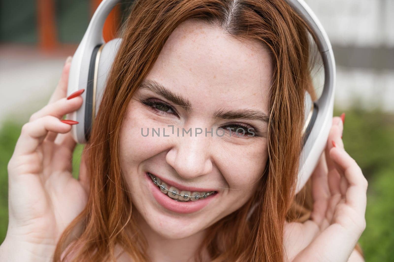 Portrait of a young red-haired woman with braces on her teeth listening to music on headphones outdoors. by mrwed54