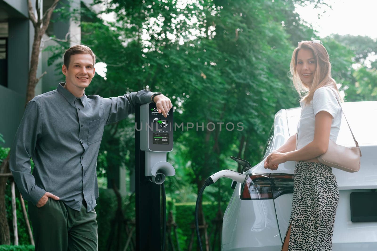 Young couple travel with EV electric car charging in green sustainable city outdoor garden in summer shows urban sustainability lifestyle by green clean rechargeable energy of electric vehicle innards