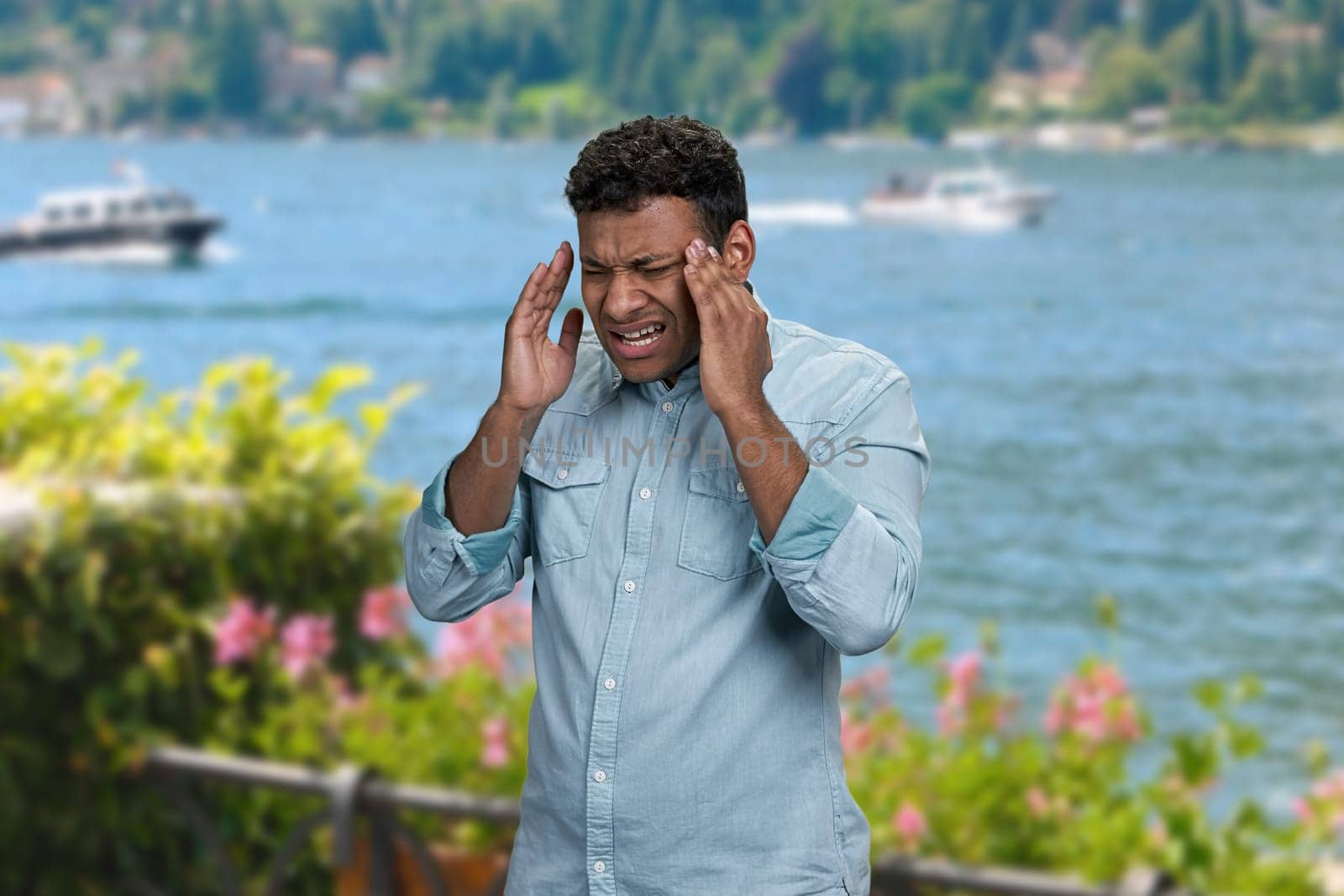 Stressed young man suffering from terrible headache standing outdoors. Man with headache on sea background.