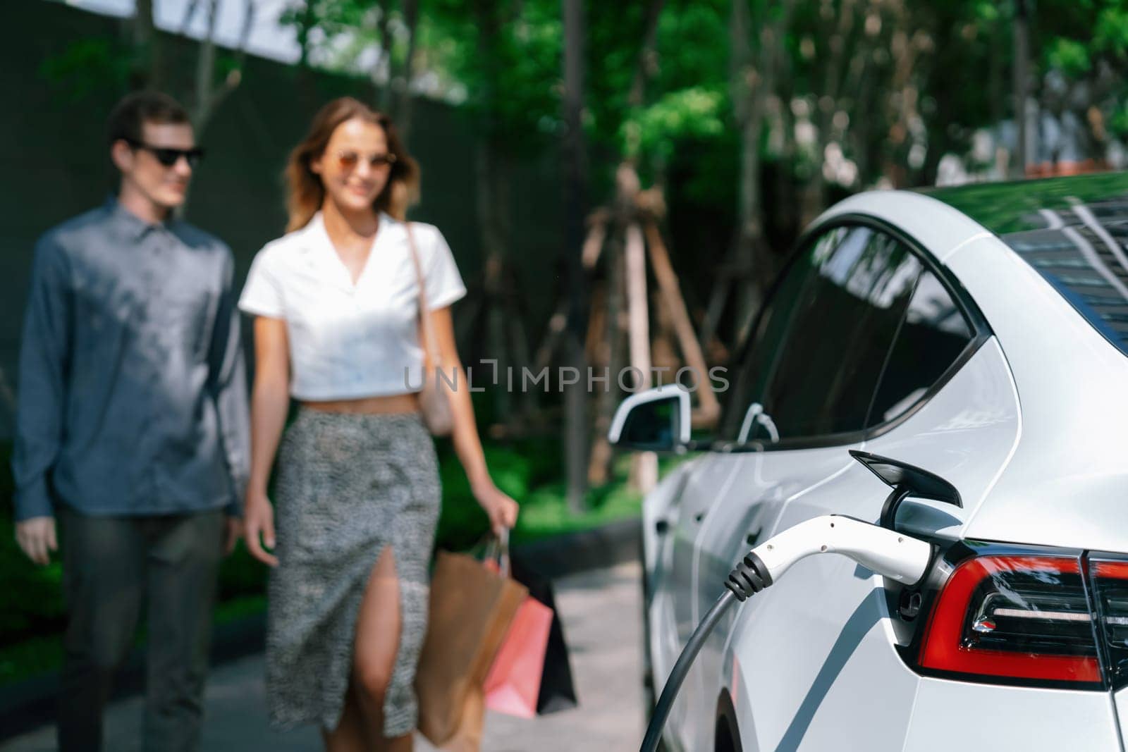 Young couple travel with EV electric car charging in green sustainable city outdoor garden in summer shows urban sustainability lifestyle by green clean rechargeable energy of electric vehicle innards