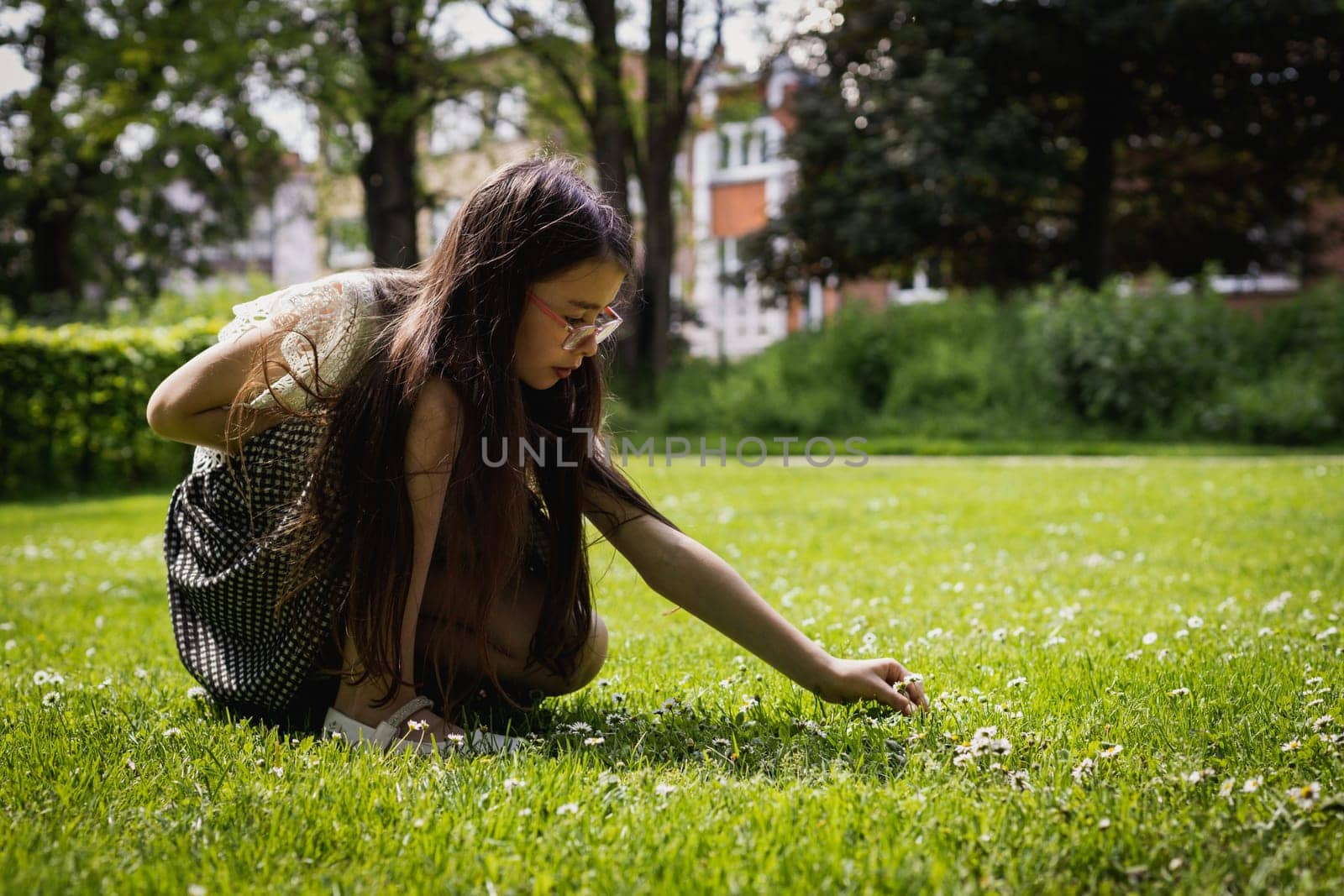 Portrait of one beautiful Caucasian brunette girl with long flowing hair, stretching out her hand and picking meadow daisies while squatting on a summer day in a public park, close-up side view.