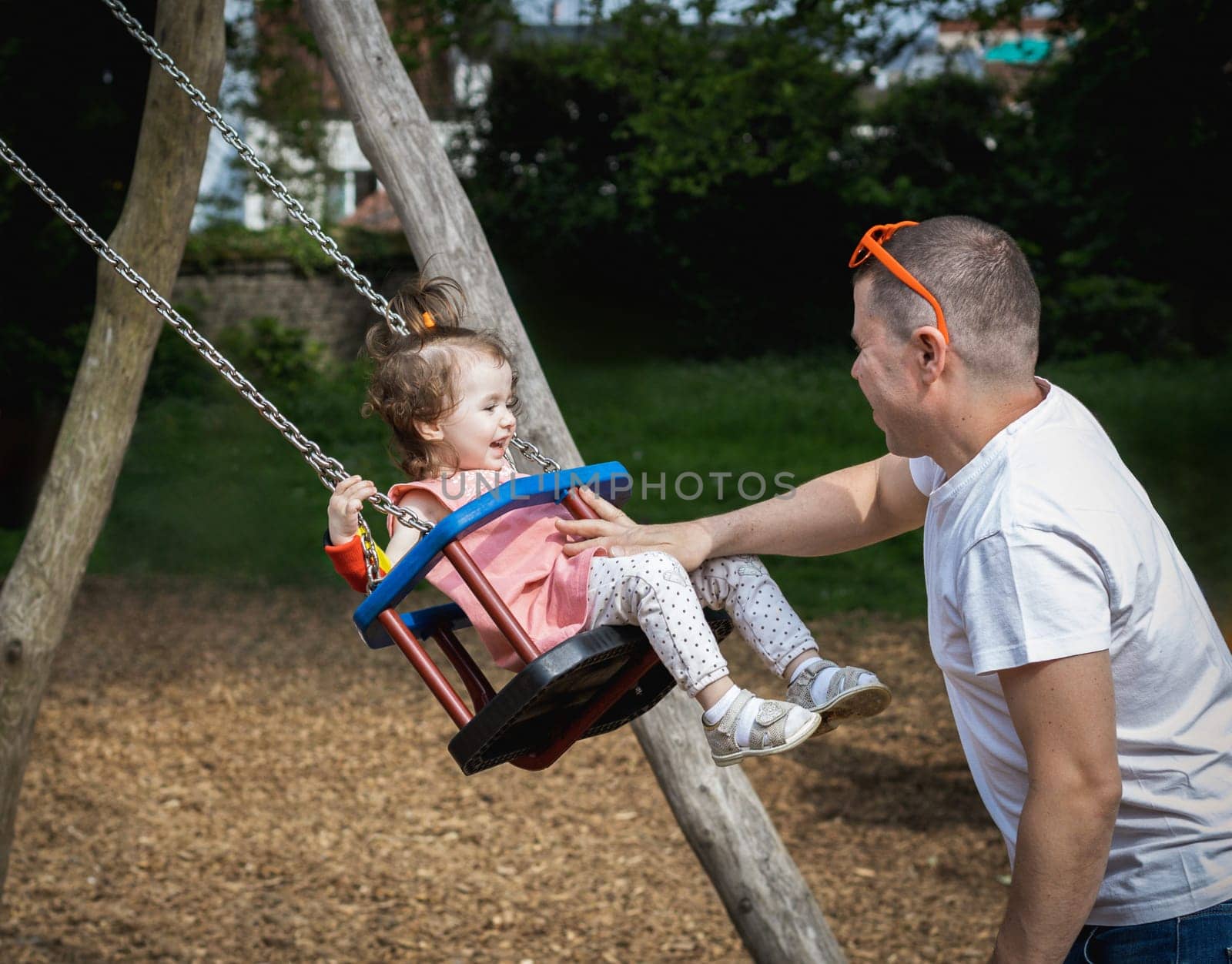 One handsome young Caucasian man father rides his little and happy laughing daughter girl on a swing on a summer day in the park on the playground, close-up side view.