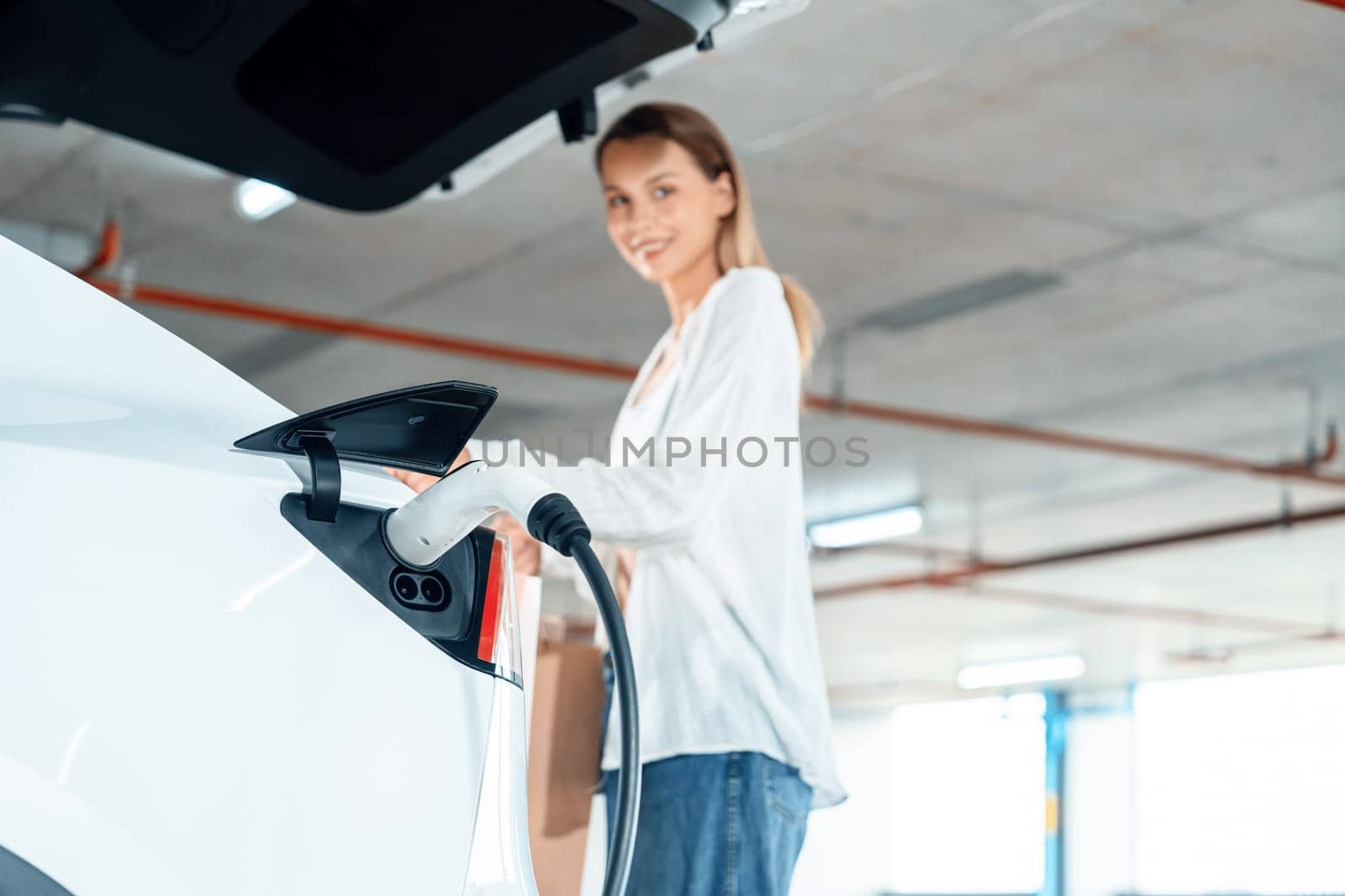 Young woman travel with EV electric car to shopping center parking lot charging in downtown city showing urban sustainability lifestyle by green clean rechargeable energy of electric vehicle innards