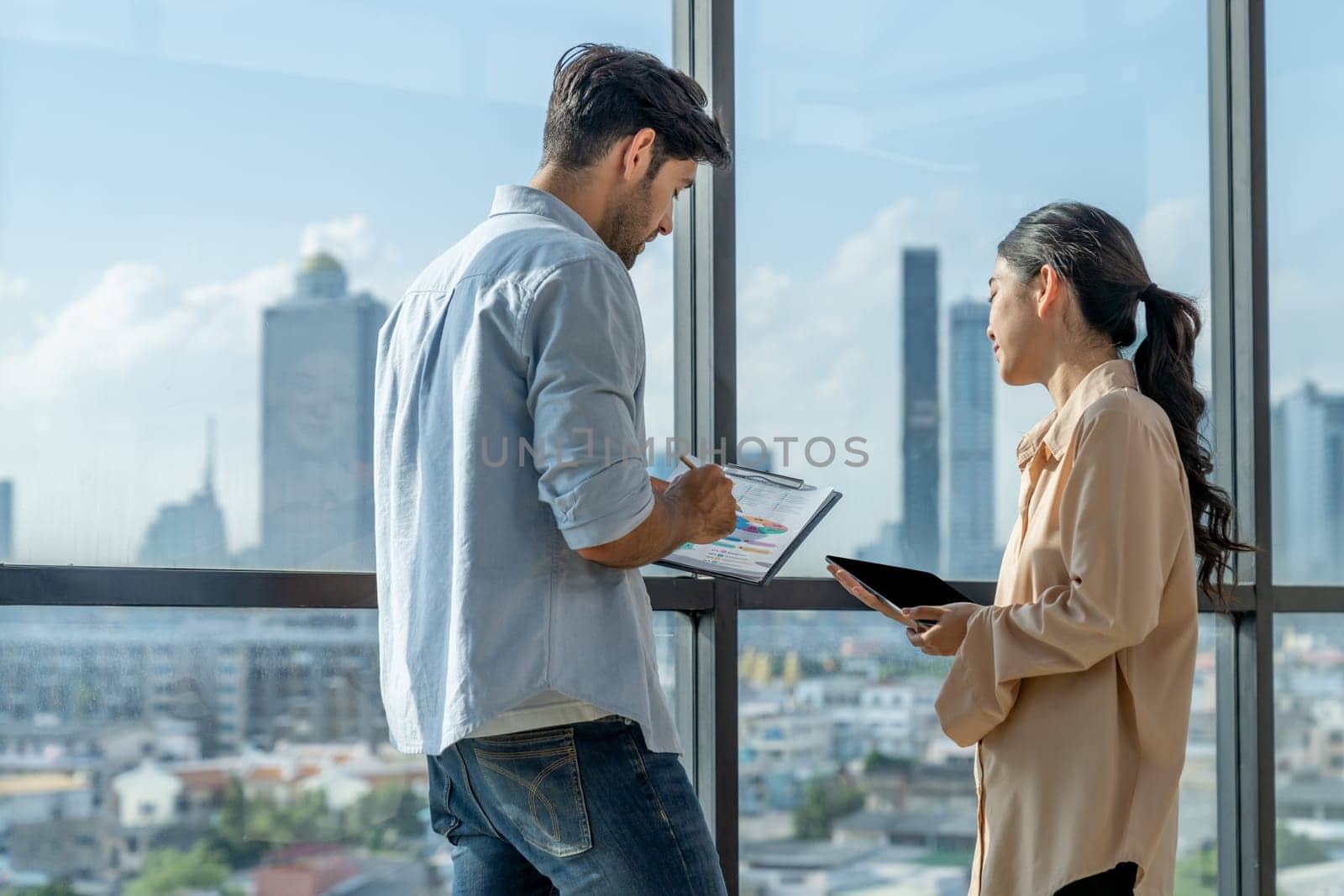 Professional businessman present financial statistic to investor while standing near panoramic window shows skyscraper or city view. Diverse businesspeople discuss about marketing plan. Tracery.