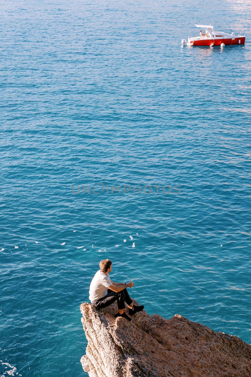 Young man sits on a rock ledge and looks at a boat sailing on the sea. Back view by Nadtochiy