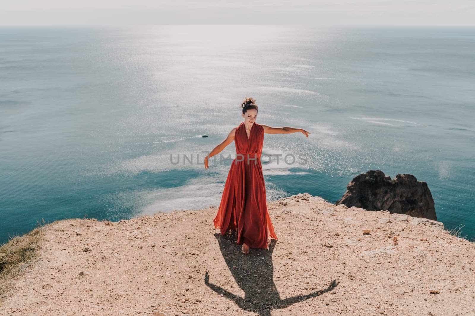 Woman red dress sea. posing on a rocky outcrop high above the sea. Girl on the nature on blue sky background. Fashion photo. by Matiunina