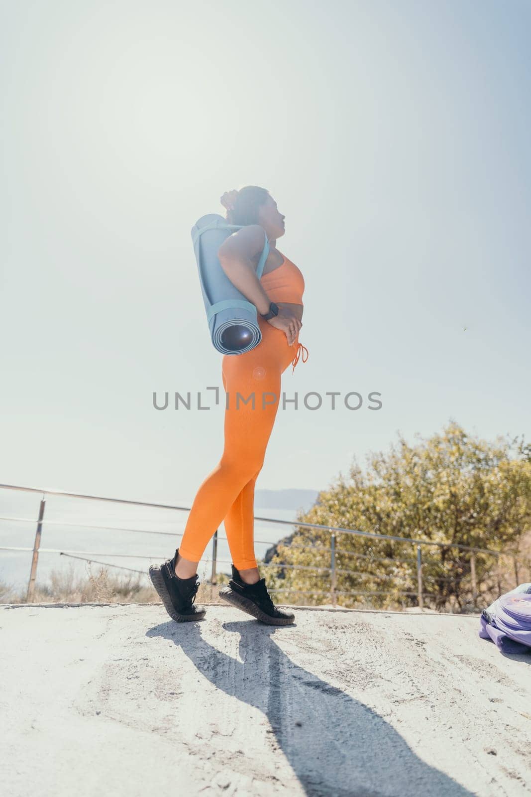 Fitness woman sea. Happy middle aged woman in orange sportswear exercises morning outdoors on yoga mat with laptop in park over ocean beach. Female fitness pilates yoga routine. Healthy lifestyle. by panophotograph