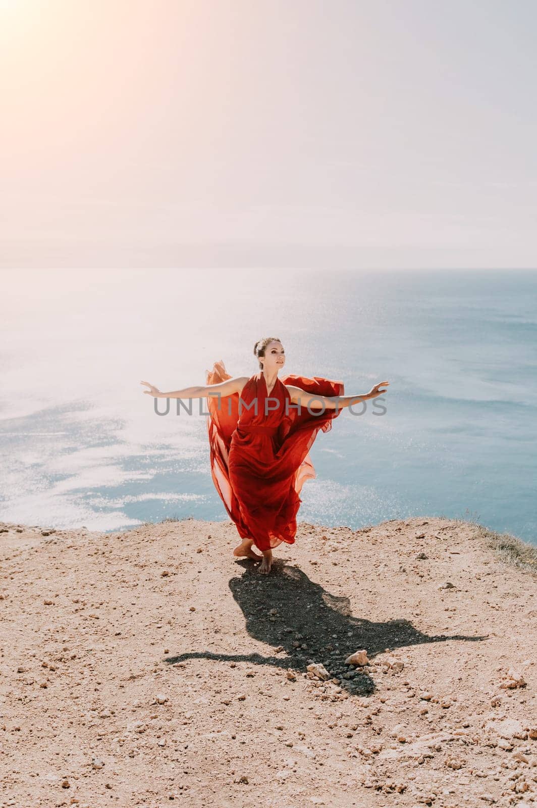 Side view a Young beautiful sensual woman in a red long dress posing on a rock high above the sea during sunrise. Girl on the nature on blue sky background. Fashion photo.