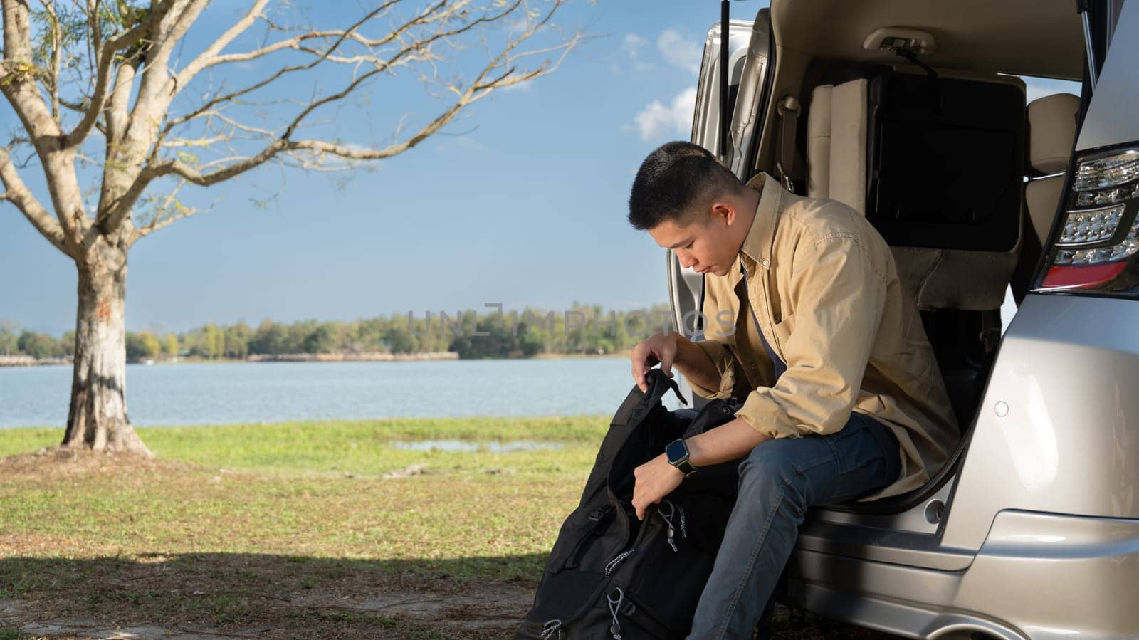 Man traveler sitting in the open trunk of minivan near near a scenic lake under the blue sky.