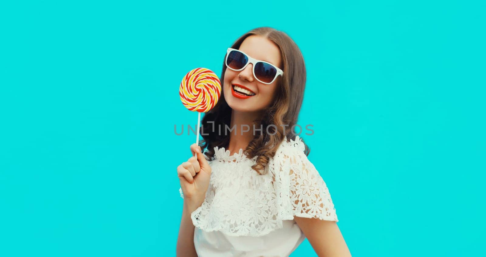 Portrait of happy caucasian smiling young woman holding colorful lollipop wearing white sunglasses on blue studio background