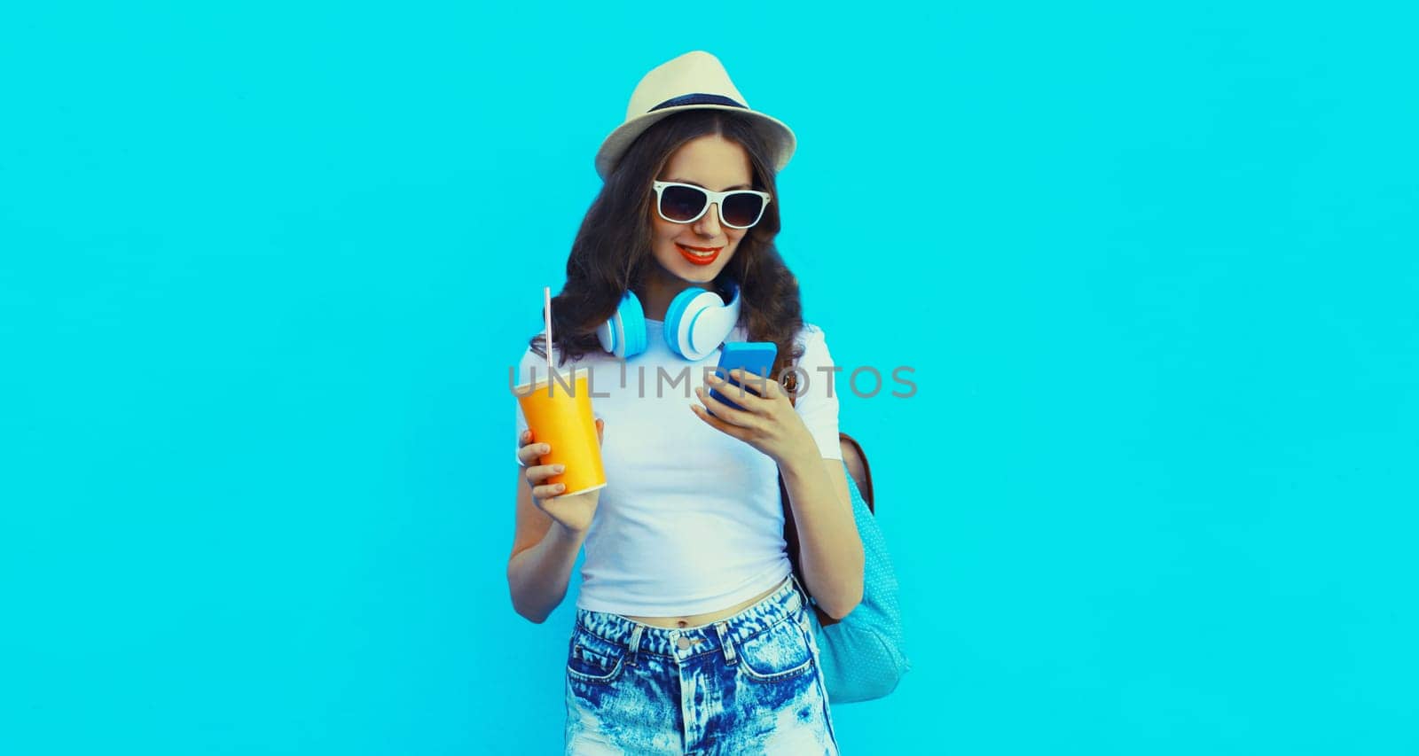 Portrait of modern caucasian young woman listening to music in headphones with mobile phone holding cup of fresh juice on blue studio background