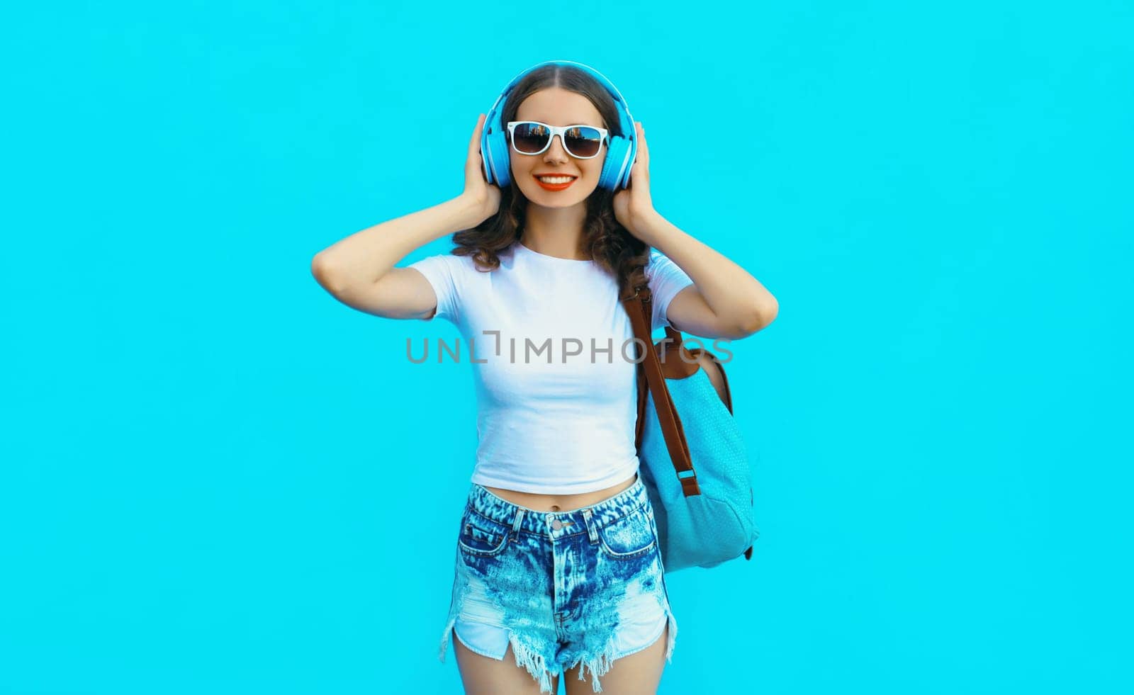 Portrait of modern happy young woman listening to music with headphones on blue studio background