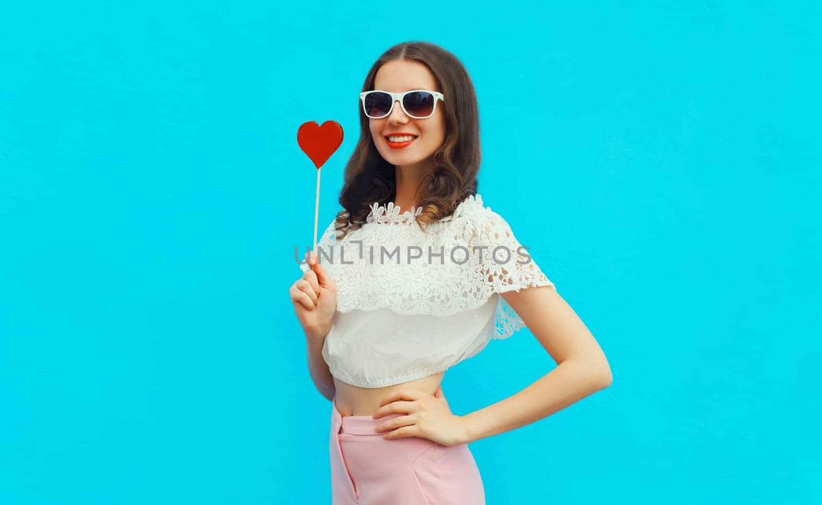 Portrait of beautiful young woman with red heart shaped lollipop on stick wearing white sunglasses on blue studio background