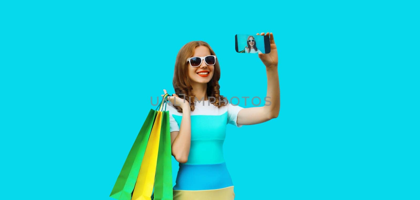 Beautiful happy smiling woman taking selfie with phone with shopping bags wearing dress on blue studio background
