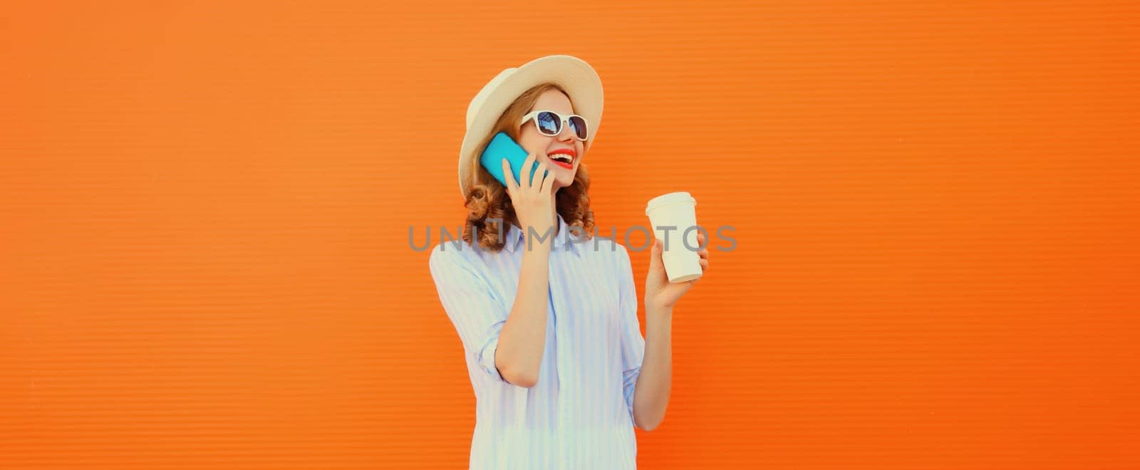 Happy cheerful smiling young woman calling on mobile phone with cup of coffee wearing summer straw hat on orange background