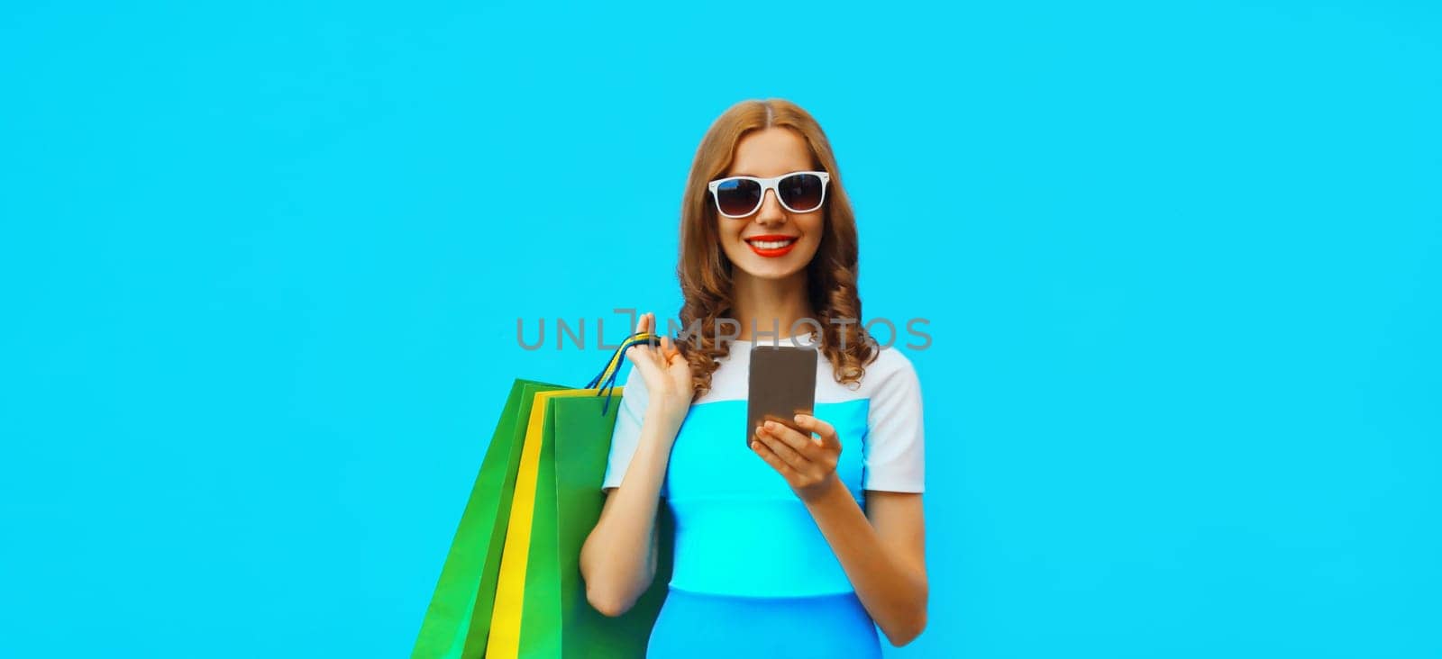 Beautiful happy young woman looking at mobile phone with shopping bag wearing summer dress on blue studio background