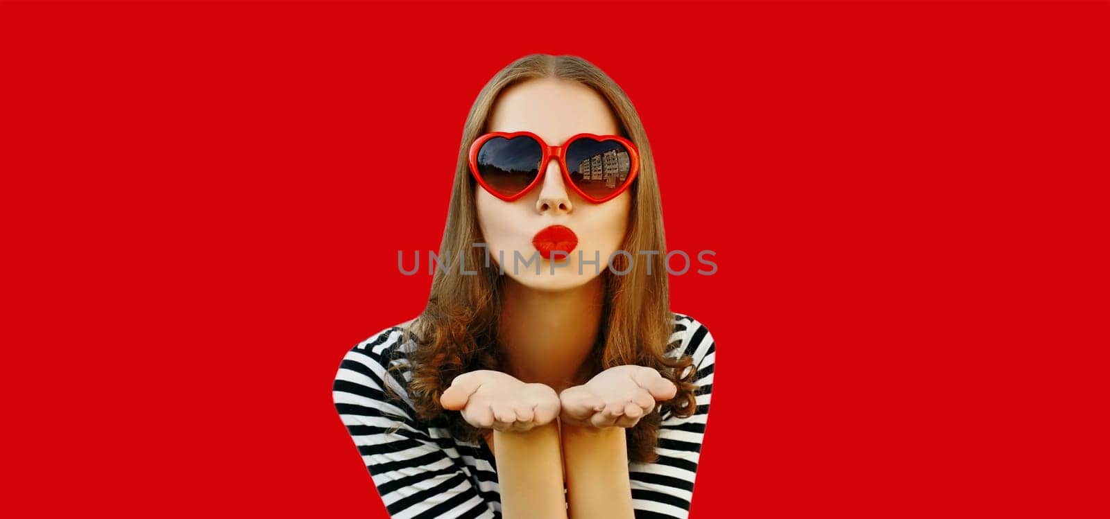 Portrait of beautiful young woman blowing her lips with red lipstick sends sweet air kiss wearing heart shaped sunglasses on studio background