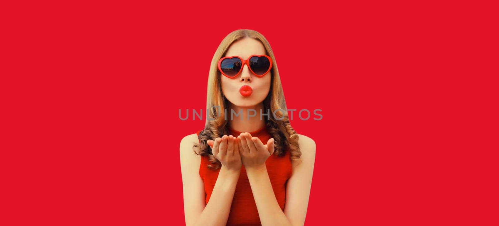 Portrait of beautiful young woman blowing her lips with red lipstick sends sweet air kiss wearing heart shaped sunglasses on studio background