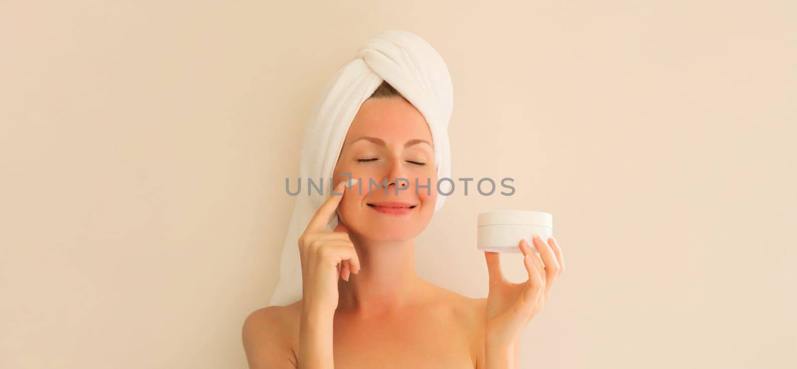 Natural beauty portrait of happy smiling young caucasian woman touches her clean skin applying face cream while drying wet hair with white wrapped bath towel on her head