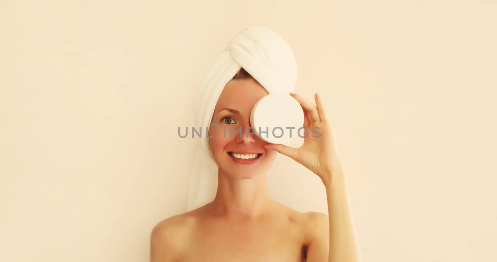 Natural beauty portrait of happy smiling young caucasian woman touches her clean skin applying face cream while drying wet hair with white wrapped bath towel on her head