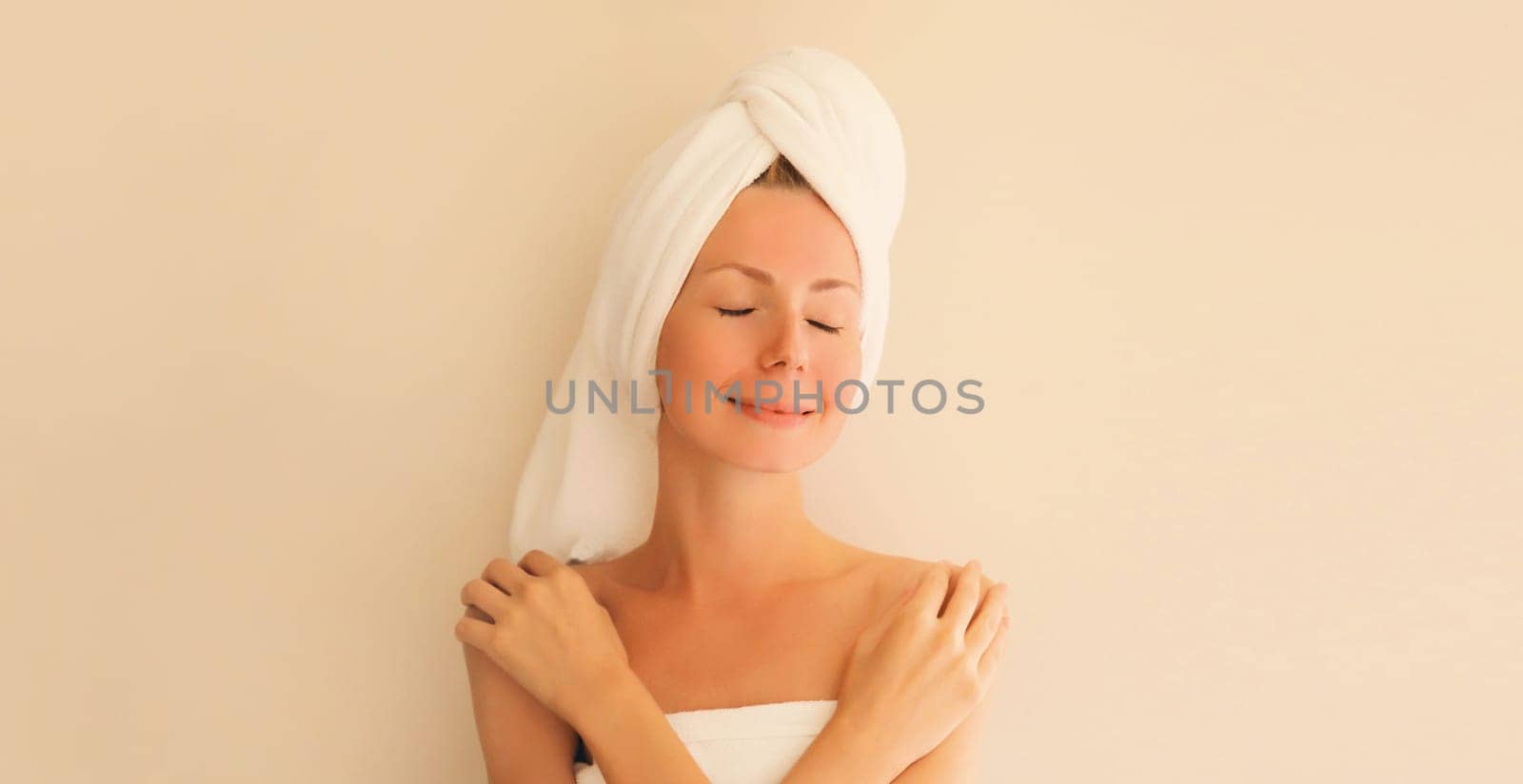 Natural beauty portrait of happy smiling young caucasian woman touches her clean skin while drying wet hair with white wrapped bath towel on her head