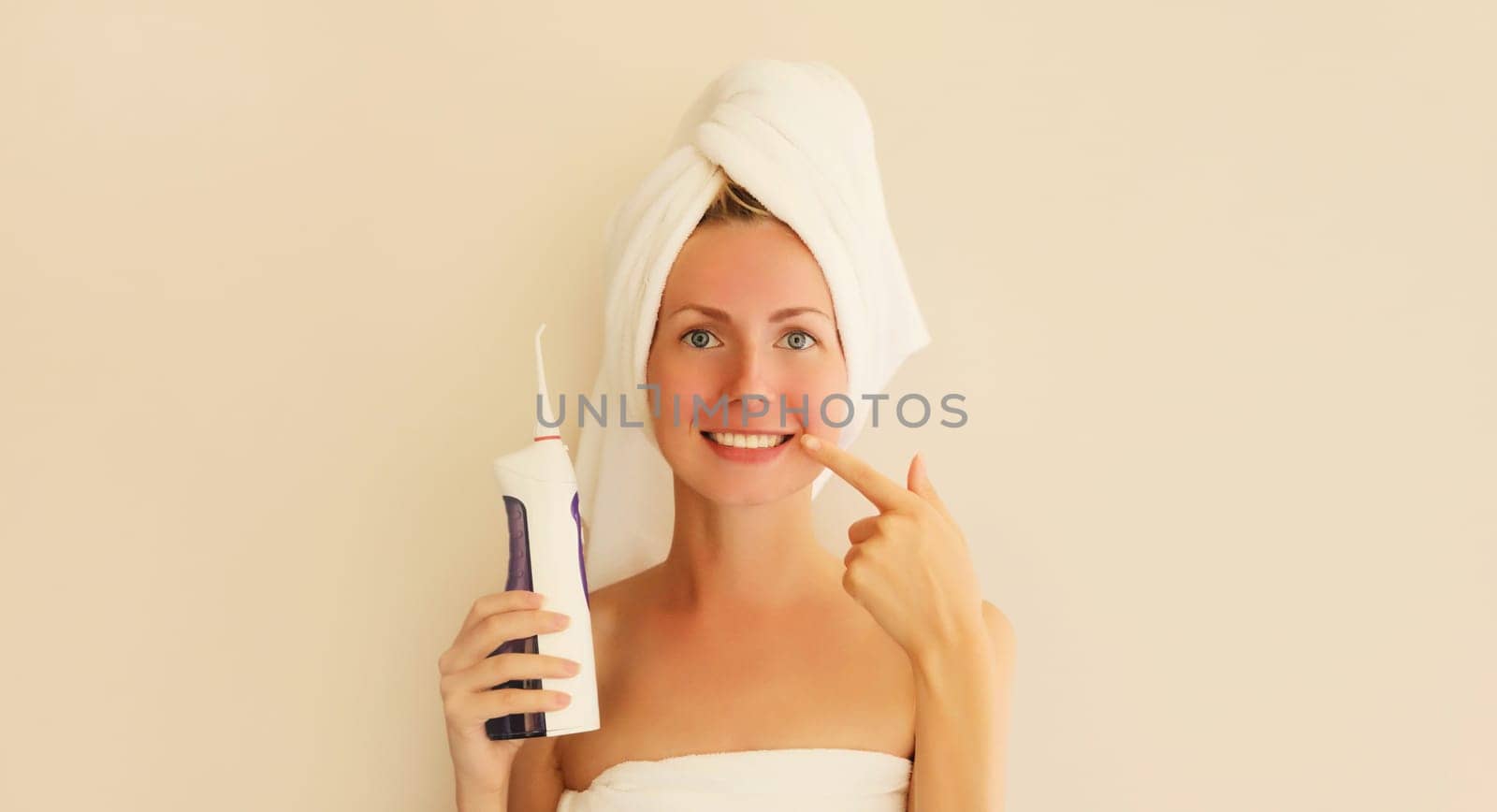 Happy smiling woman points to her clean white teeth holds oral irrigator with bath towel on head by Rohappy