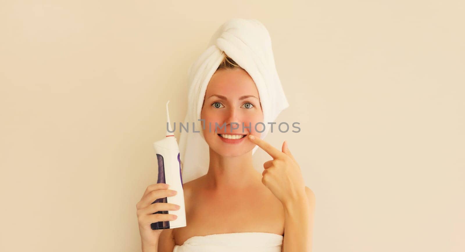 Portrait of happy smiling young woman points to her clean white teeth holds oral irrigator and dries her wet hair with wrapped towel on her head in the morning at home