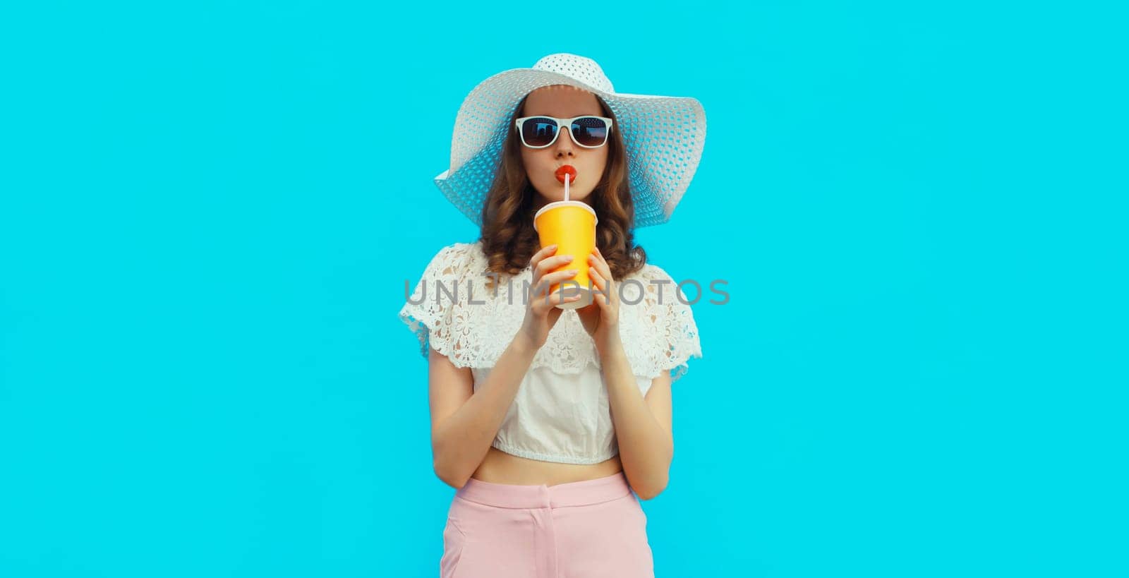 Portrait of beautiful caucasian young woman model with cup of coffee or fresh juice wearing white summer straw hat on studio blue background