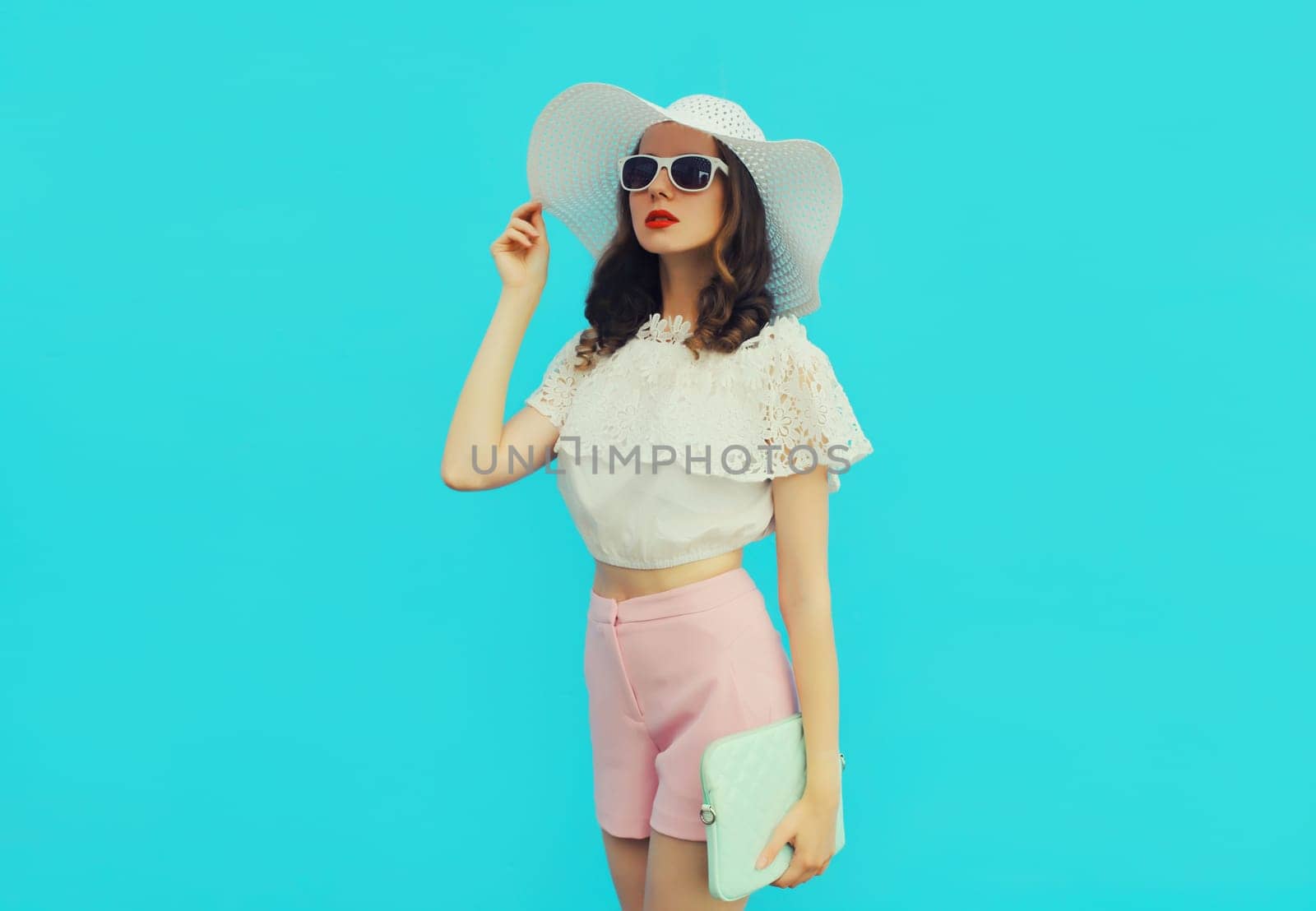 Beautiful caucasian young woman model posing with handbag clutch wearing white summer straw hat on blue studio background