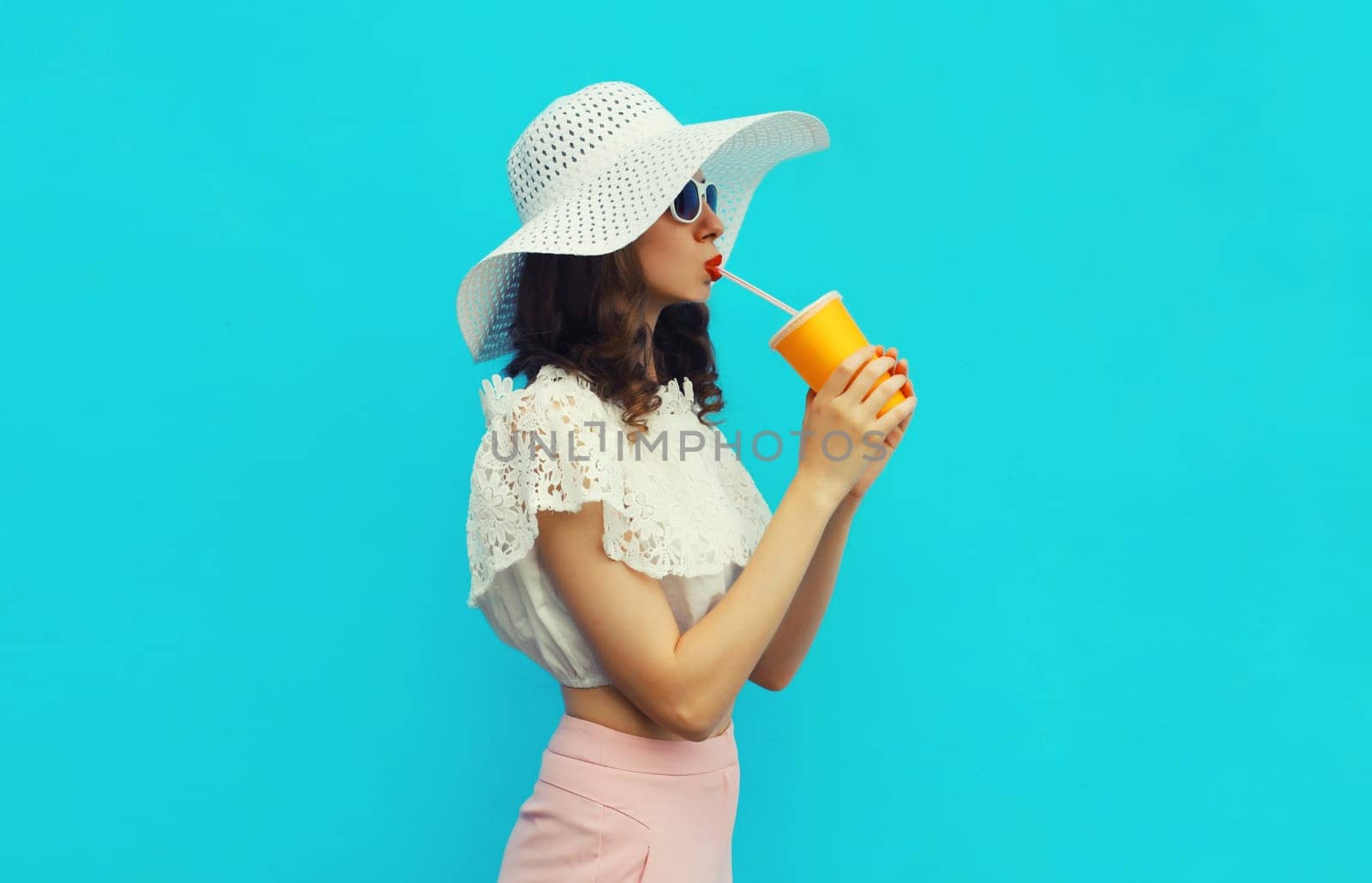 Portrait of beautiful caucasian young woman model with cup of coffee or fresh juice wearing white summer straw hat on studio blue background