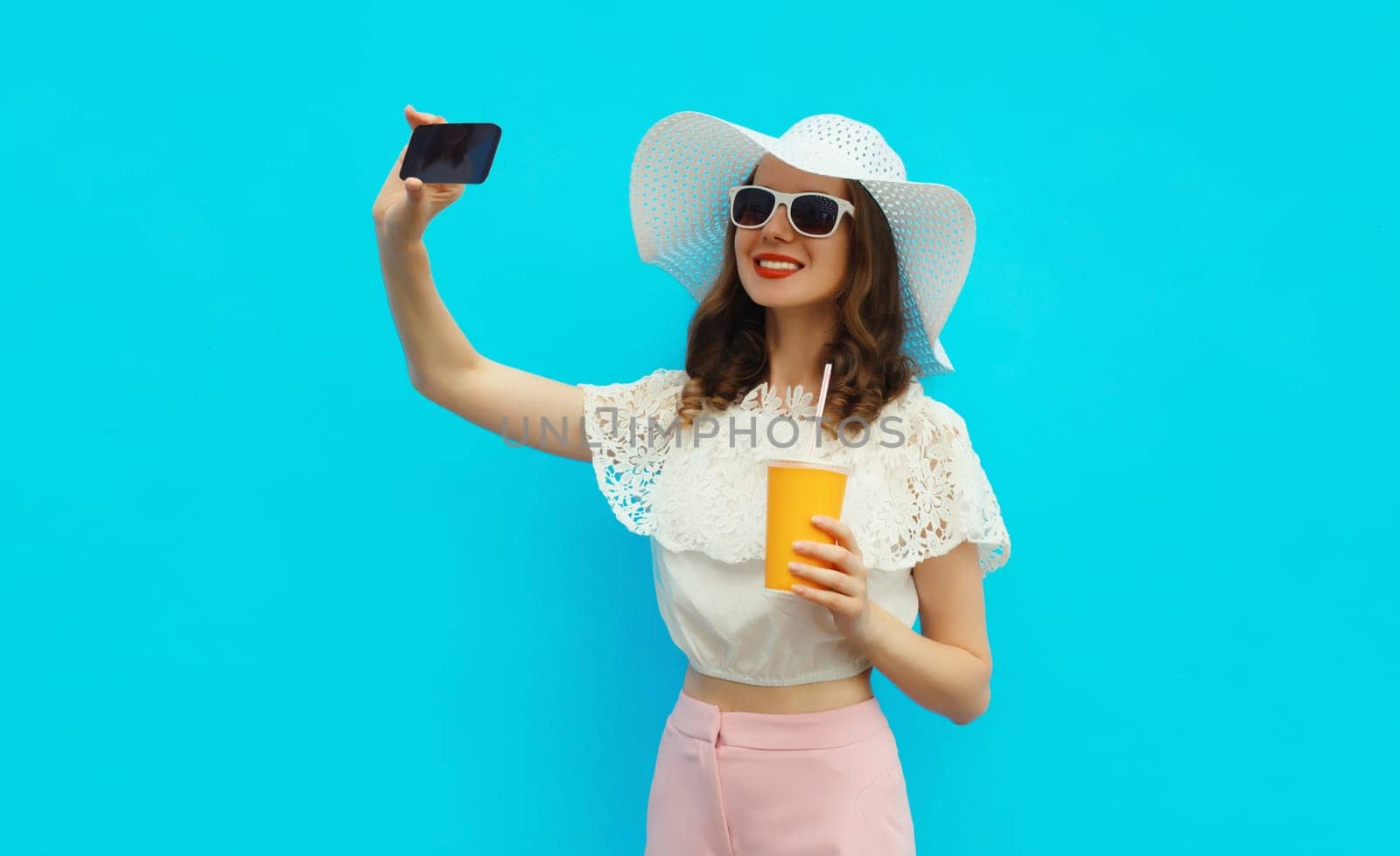 Summer portrait of beautiful happy smiling young woman taking selfie with mobile phone and cup of fresh juice wearing white straw hat on colorful blue background
