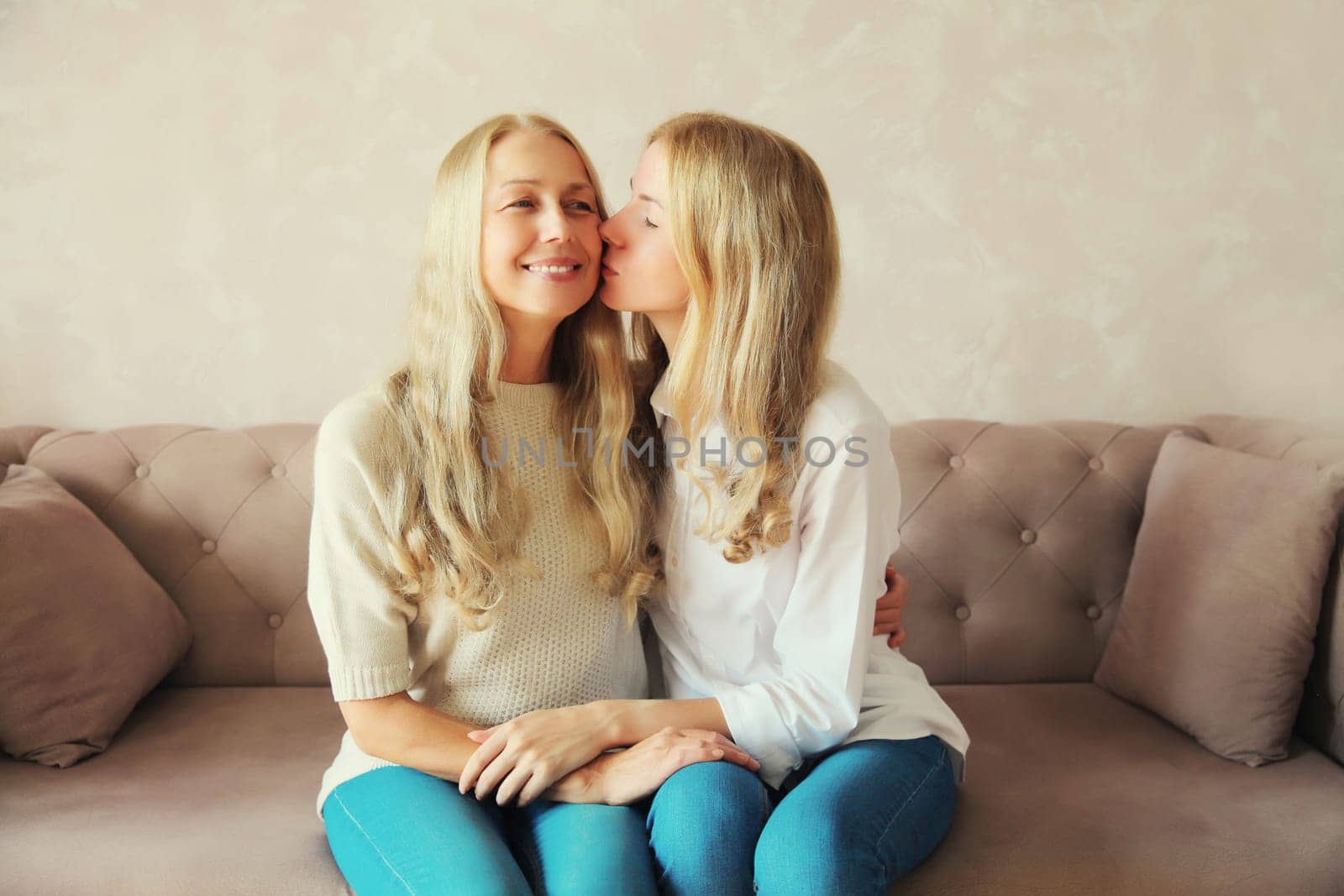 Portrait of caucasian adult daughter kissing her happy smiling middle-aged mother while sitting on couch at home