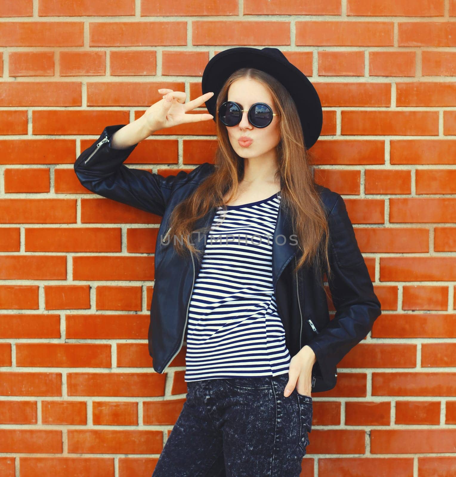 Portrait of stylish young woman model wearing black round hat, leather jacket in rock style on city street on brick wall background