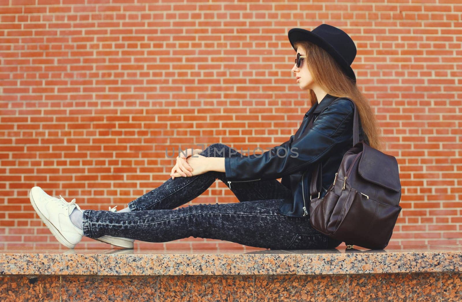 Portrait of stylish young woman model wearing black round hat, leather jacket in rock style on city street on brick wall background