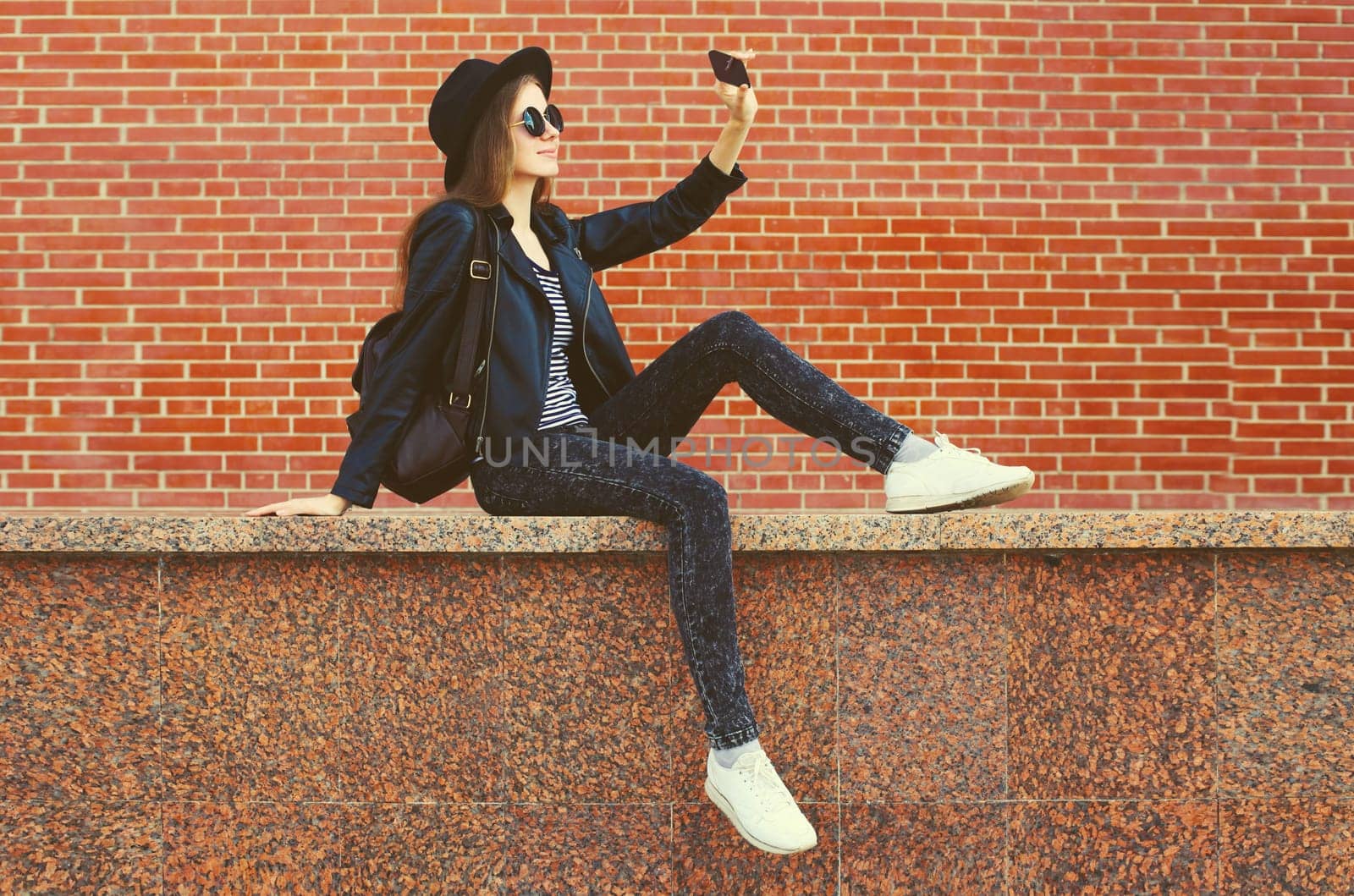 Stylish modern caucasian young woman taking selfie with mobile phone wearing black round hat, jacket and backpack on city street background