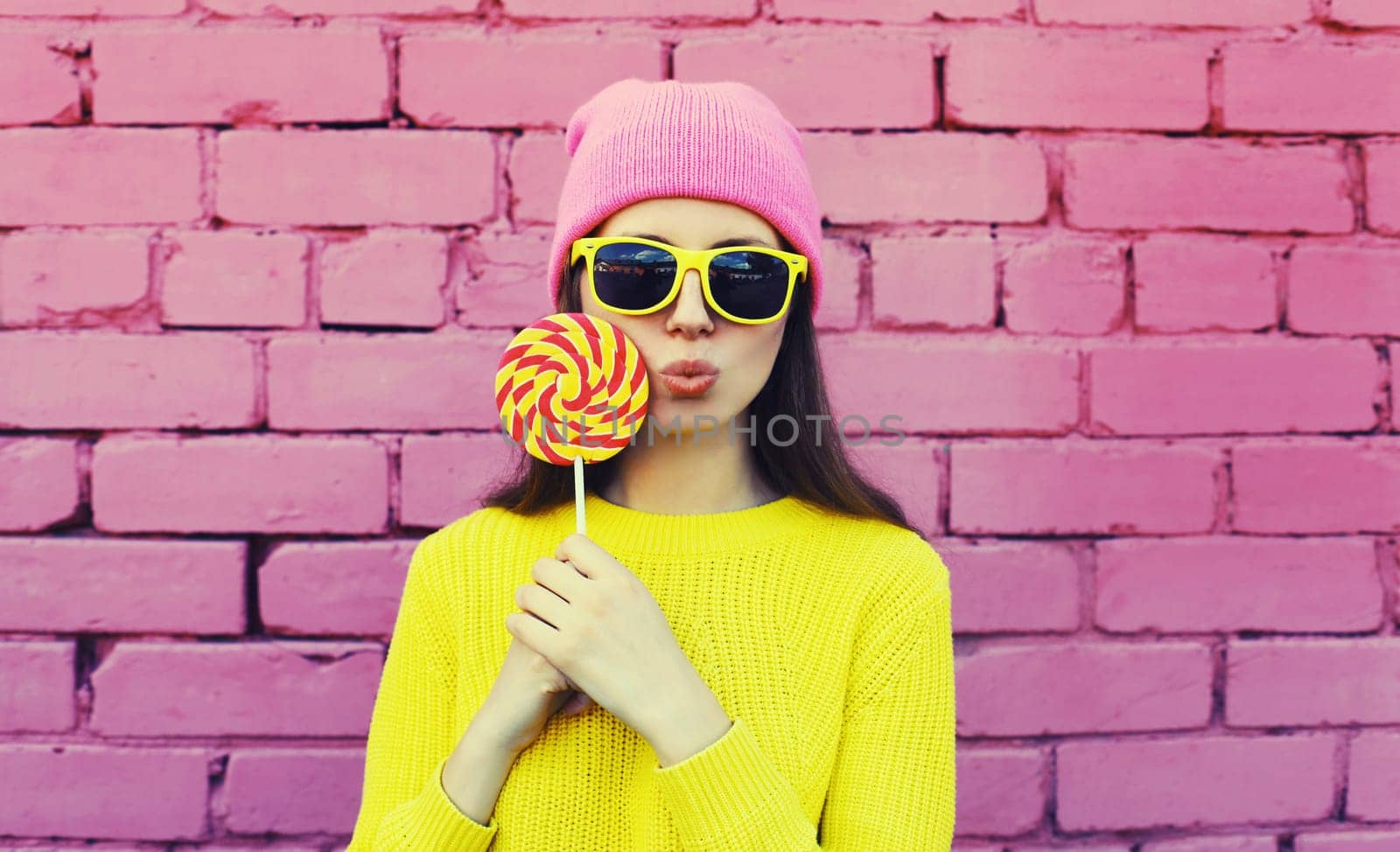 Colorful portrait of stylish modern young woman with lollipop blowing her lips sending sweet air kiss wearing vivid yellow sweater, sunglasses and pink hat on brick wall background