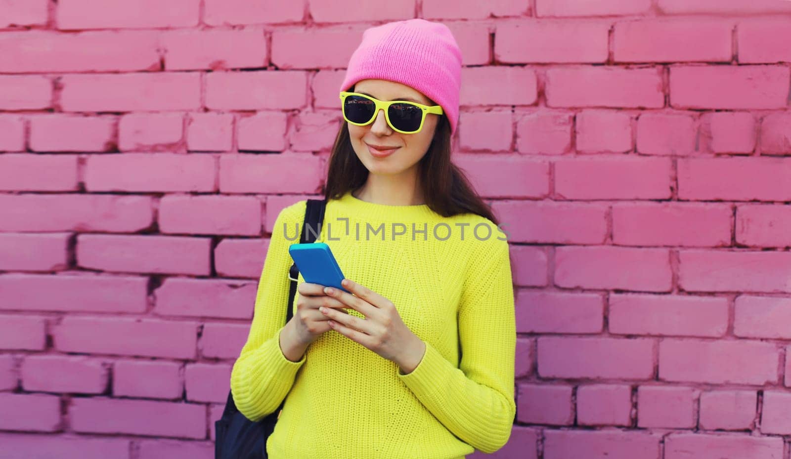 Modern caucasian young woman 20s model with mobile phone looking at device wearing colorful vivid clothes, yellow sweater and pink hat on city street