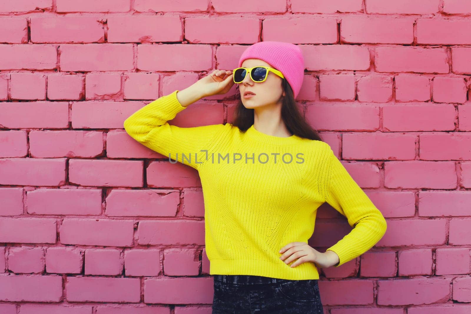 Portrait of stylish young teenager girl wearing vivid colorful clothes, knitted yellow sweater and pink hat on brick wall background